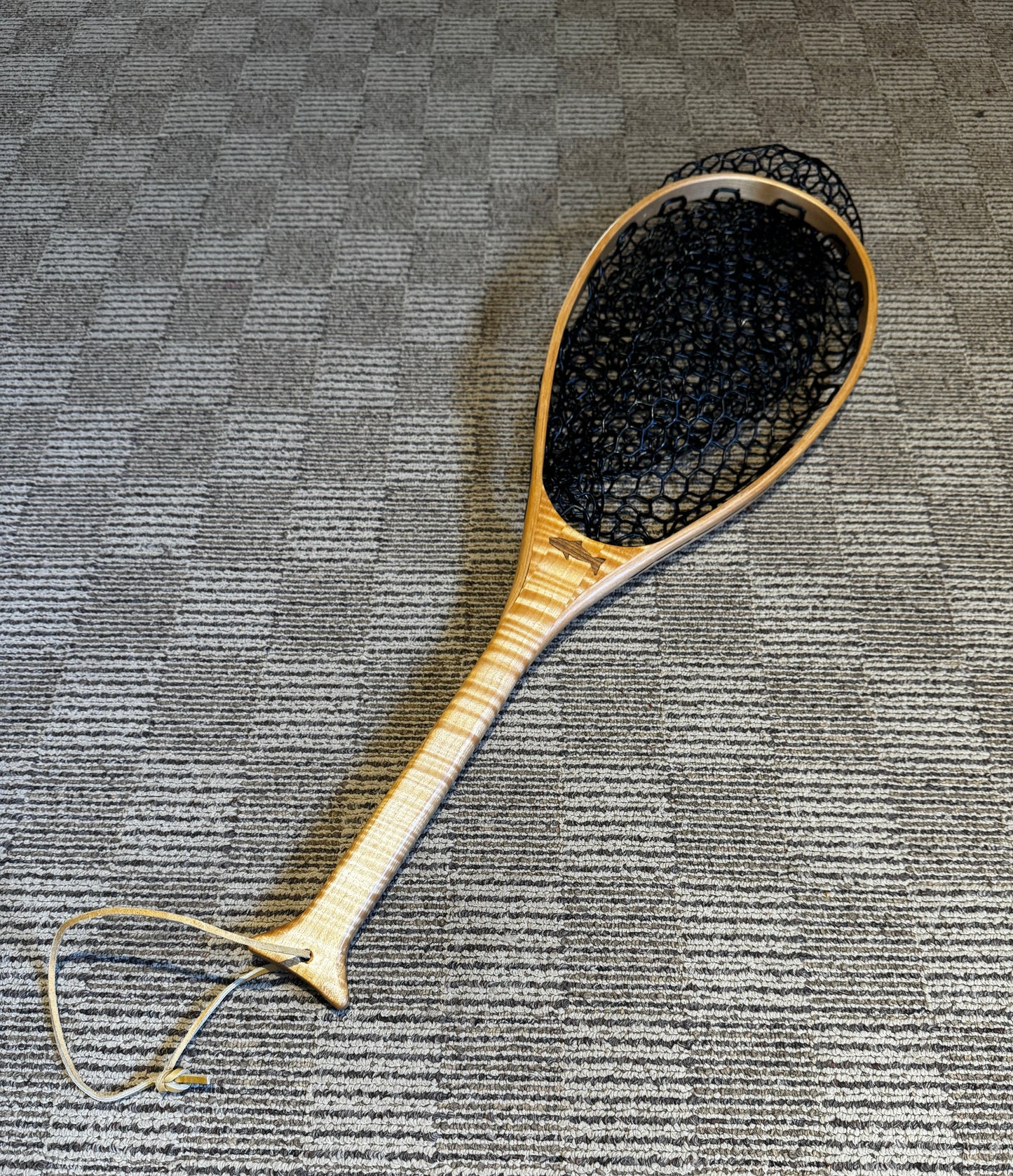 A handmade maple wood fishing net with a black rubber mesh bag, lying on a textured grey floor.